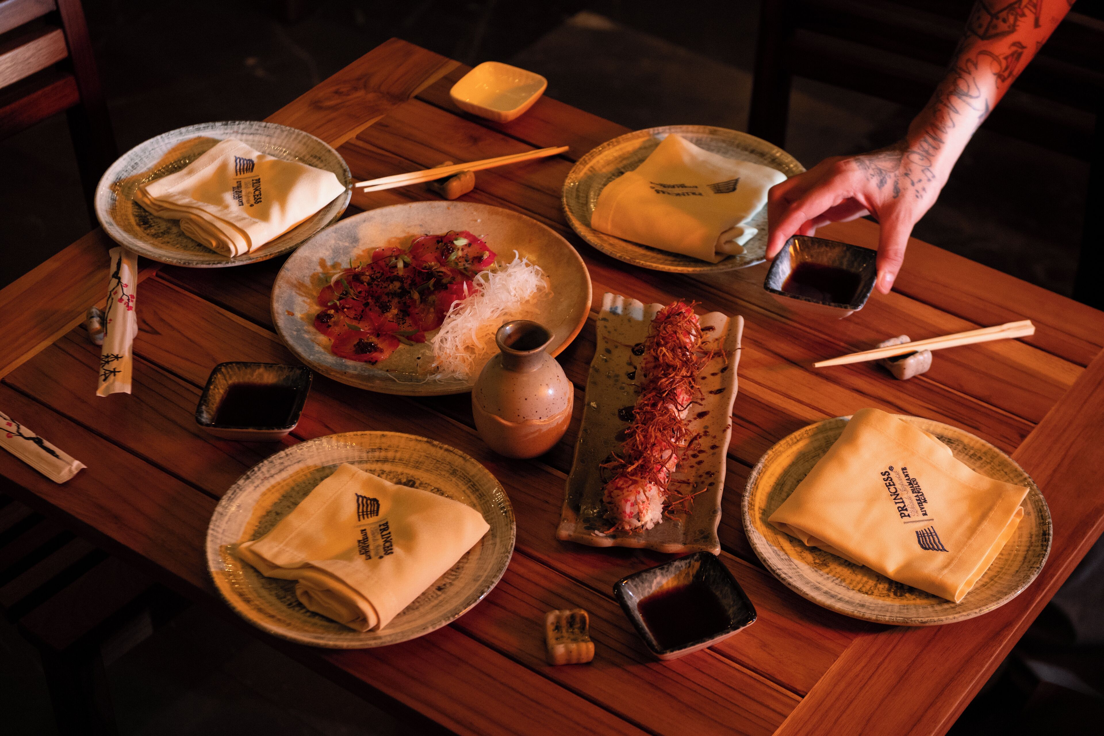 Mesa de madera con platos, palillos y una mano alcanzando un plato.