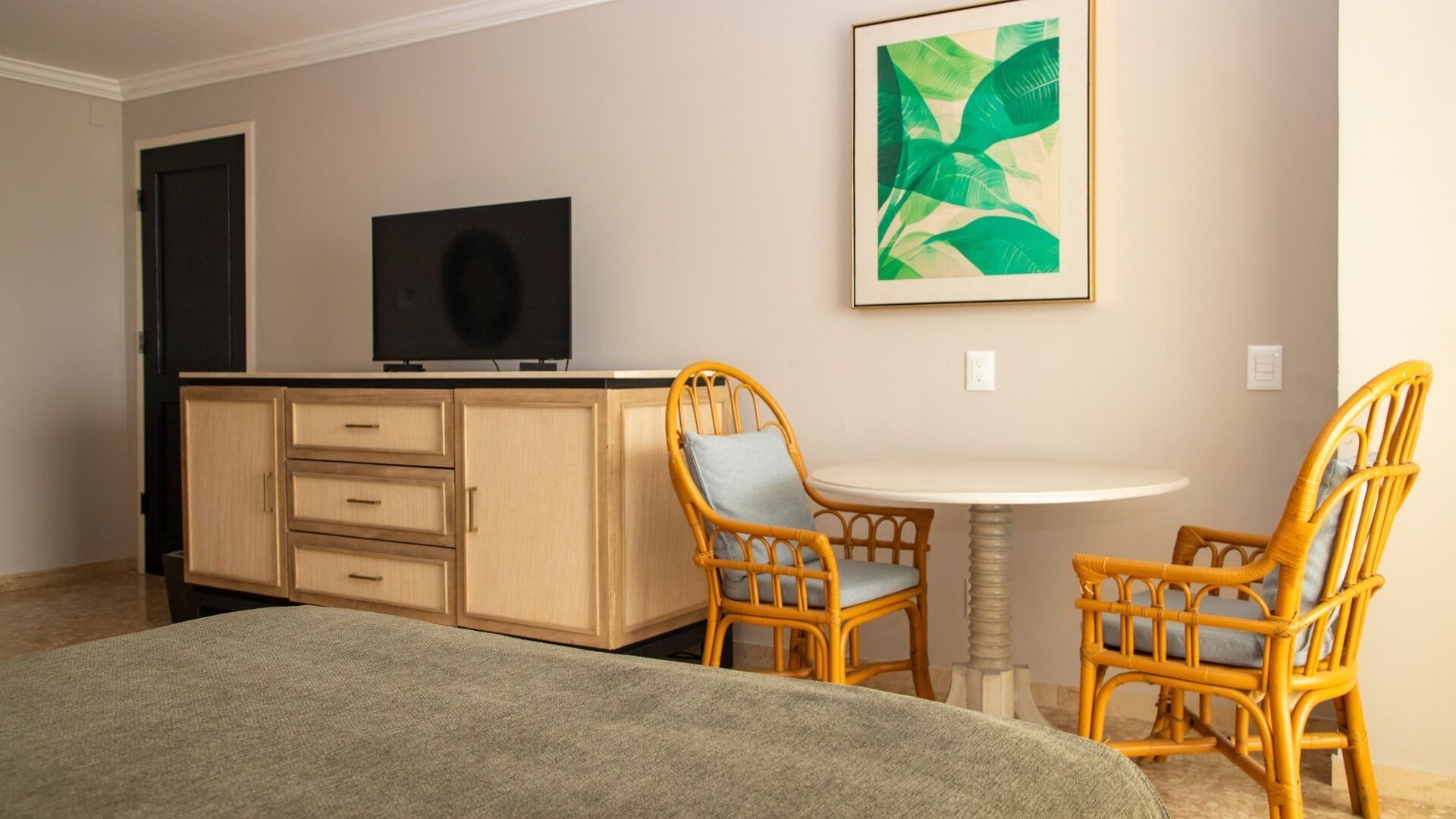Quarto com um armário de madeira, televisão, duas cadeiras de vime, uma mesa redonda e um quadro na parede.
