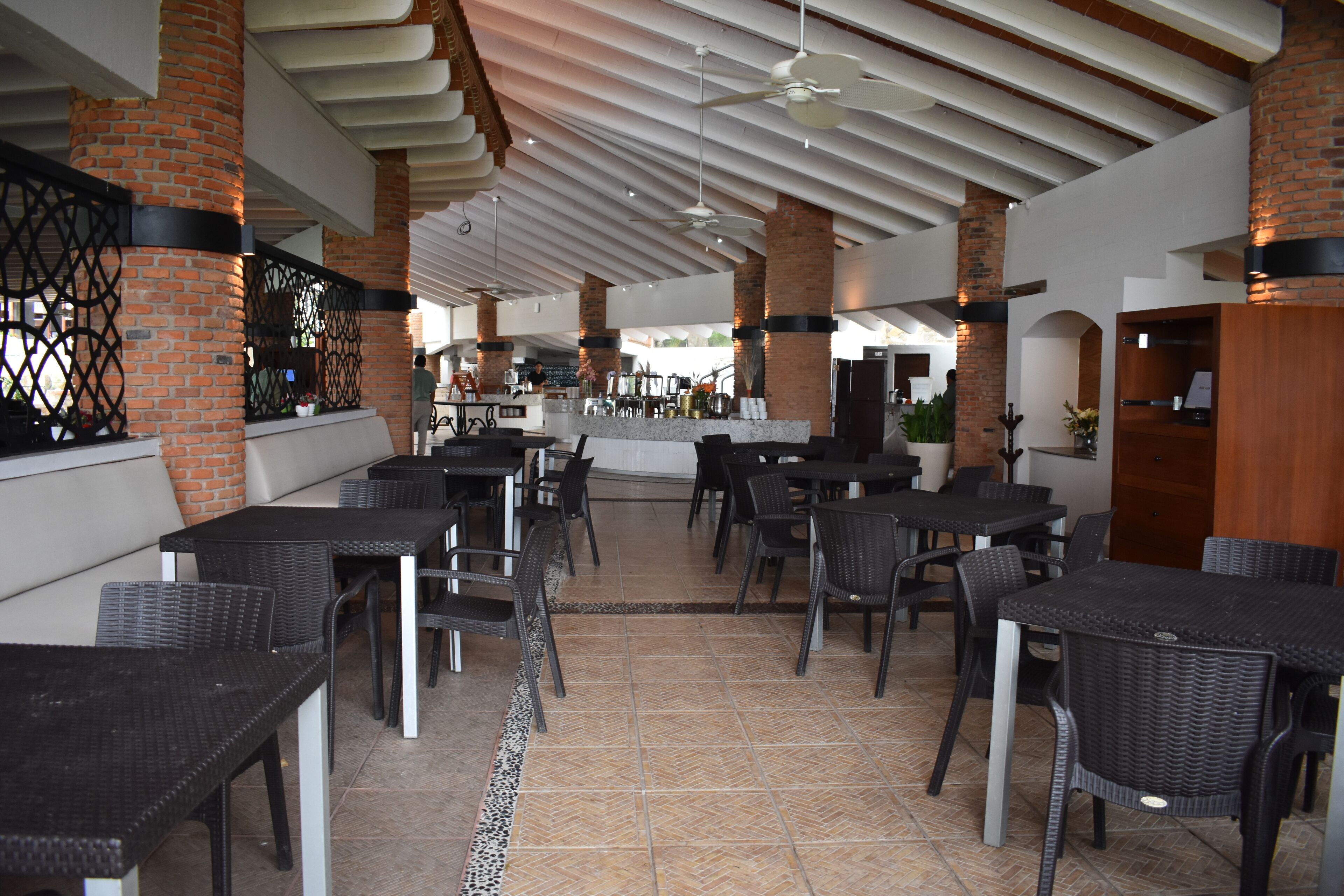Un amplio comedor interior con pilares de ladrillo, mobiliario de mimbre negro y suelo de baldosas.