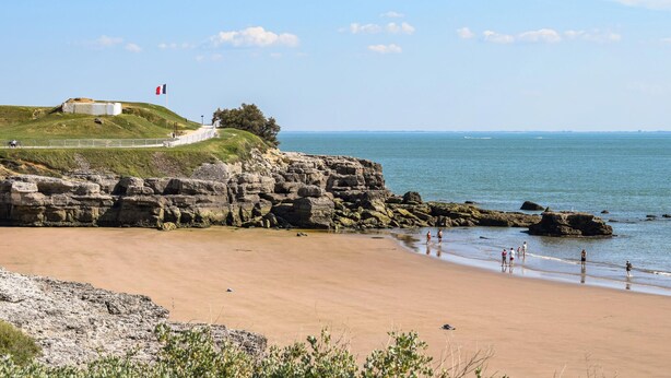 Un paisaje costero con una playa arenosa, acantilados rocosos y un pequeño edificio con una bandera en una colina cubierta de césped.