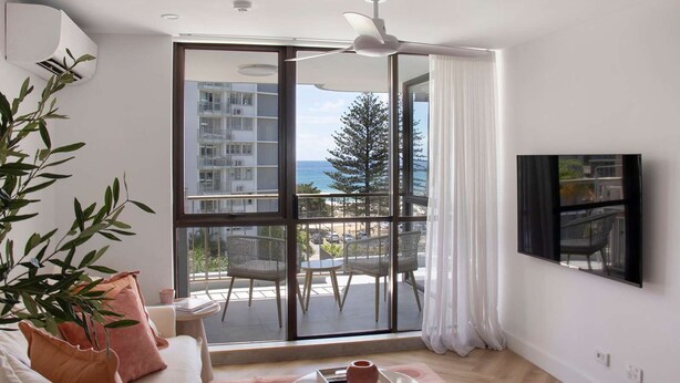 Un salon moderne doté d’une grande porte-fenêtre coulissante donnant sur un balcon, d’un ventilateur de plafond, d’un téléviseur à écran plat fixé au mur et d’une plante en pot.