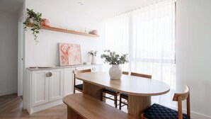 Une cuisine moderne avec des meubles blancs, une table à manger en bois et une grande fenêtre ornée de rideaux légers.