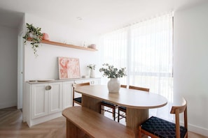 A modern kitchen with white cabinetry, a wooden dining table, and a large window with sheer curtains. - Blush by Khove (Burleigh Heads)