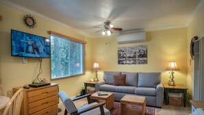 A living room with a sofa, two side tables with lamps, a wooden coffee table, a TV mounted on the wall, a ceiling fan, and a window with blinds. - Sparrow's Nest (Yosemite National Park)