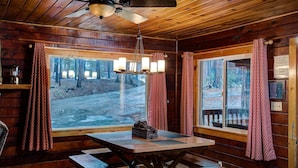 Interno di una baita con un tavolo di legno, panche, una ventola a soffitto e ampie finestre con tende.