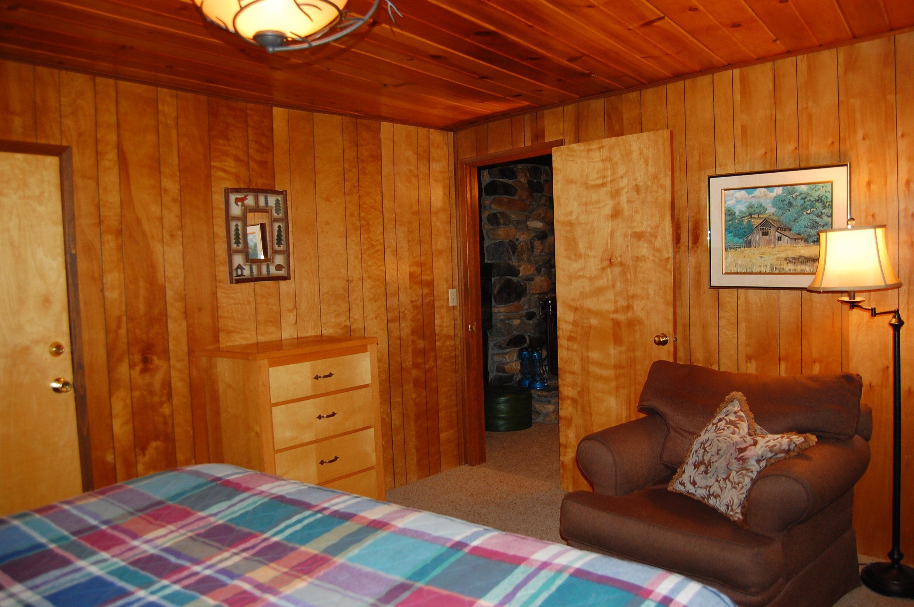 A wooden-paneled room with a bed, a dresser, a chair with a pillow, a framed picture, and a lamp.