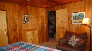 A wooden-paneled room with a bed, a dresser, a chair with a pillow, a framed picture, and a lamp.