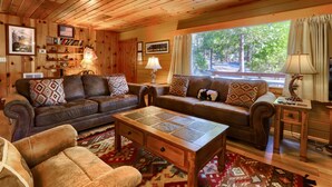 A cozy cabin-style living room with wooden walls and ceiling, a brown leather sofa, a wooden coffee table, and a large window with curtains. - Walk-a-bout (Yosemite National Park)