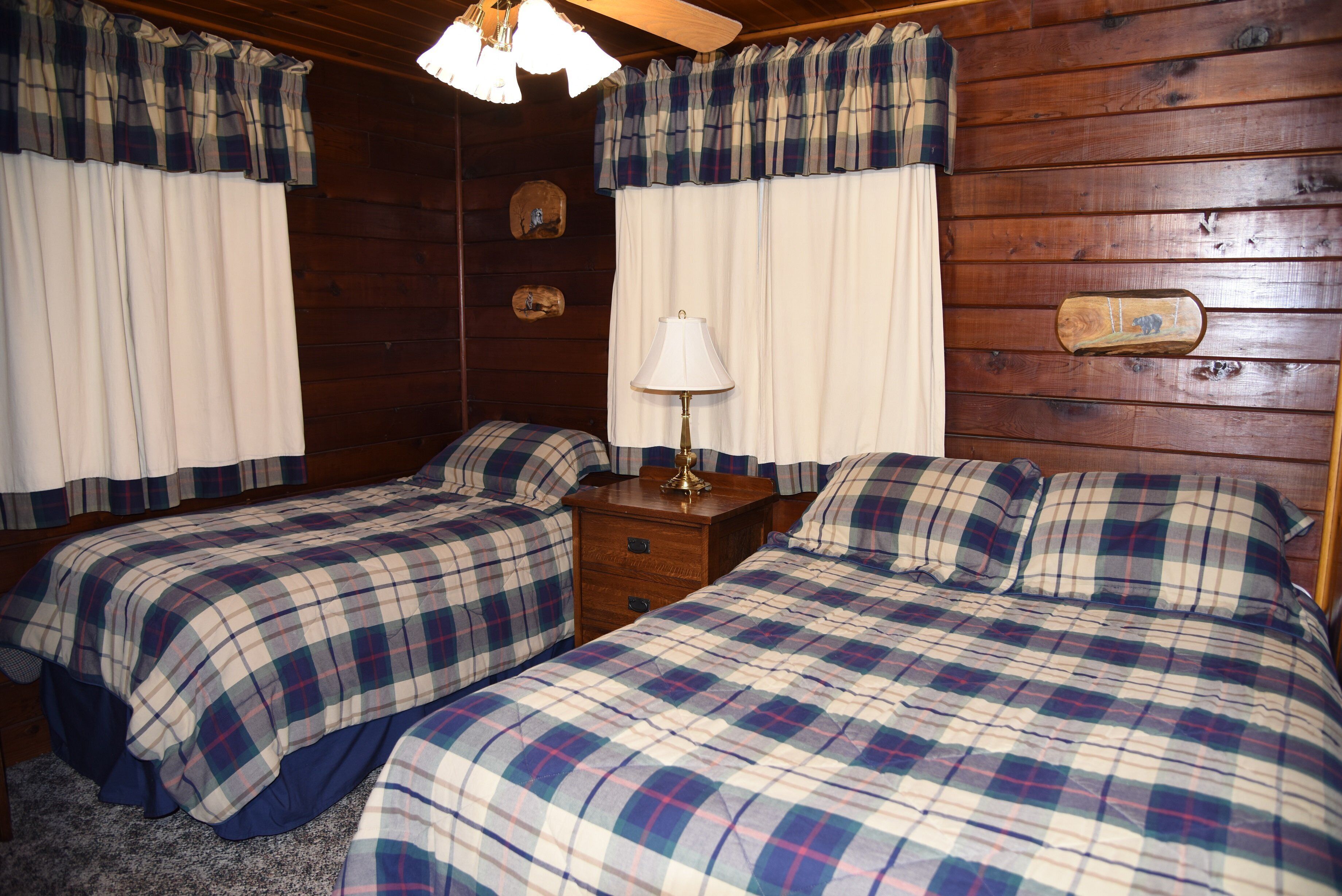 A room with two beds, each with plaid bedding, a wooden nightstand with a lamp, and wooden walls.