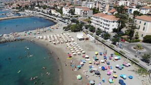 Une vue aérienne d’une plage parsemée de nombreux parasols et de chaises longues, d’un port de plaisance avec des bateaux, et de bâtiments en arrière-plan.