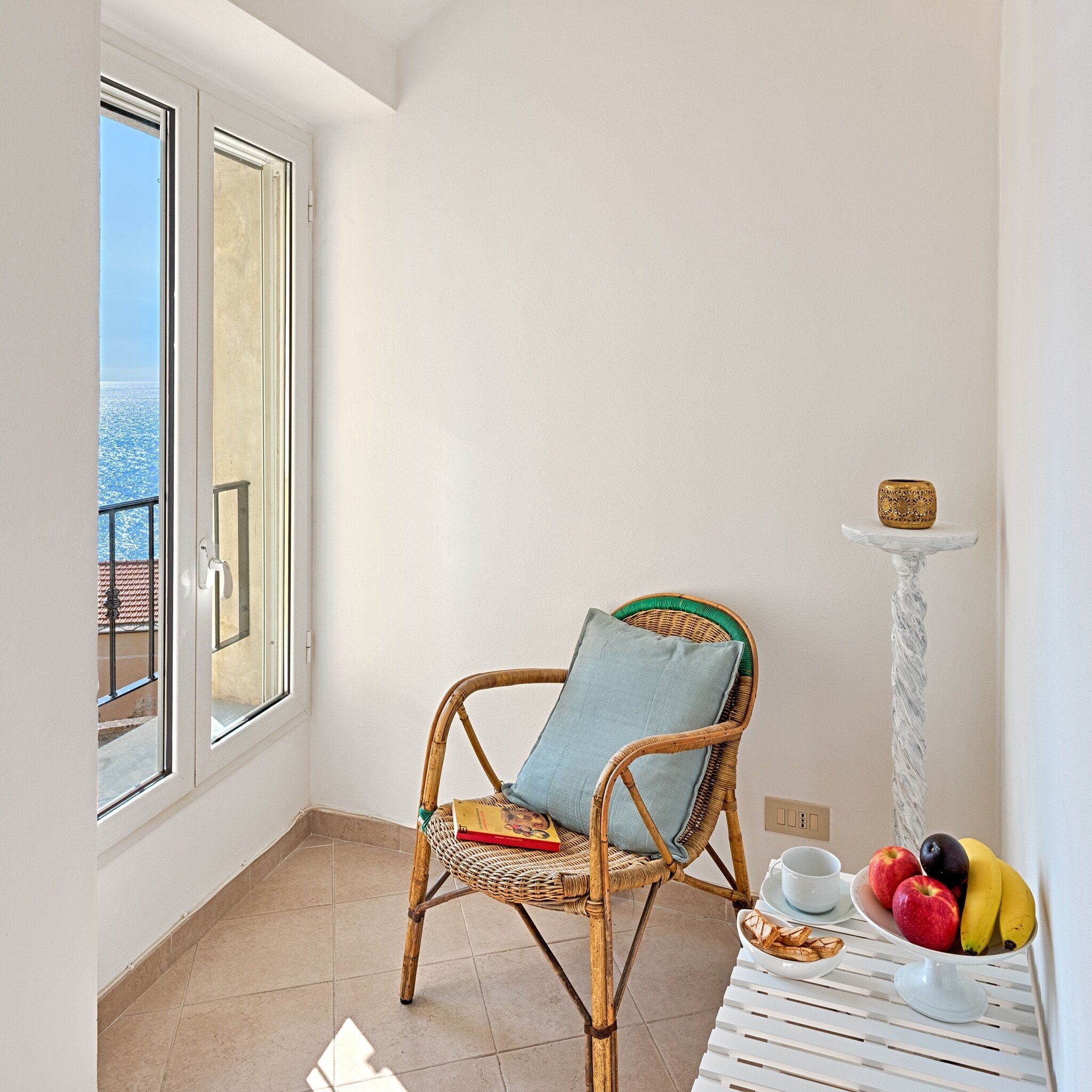 Ein Balkon mit einem Korbstuhl, einem Tisch mit Früchten und einer Tasse, und Blick aufs Meer.