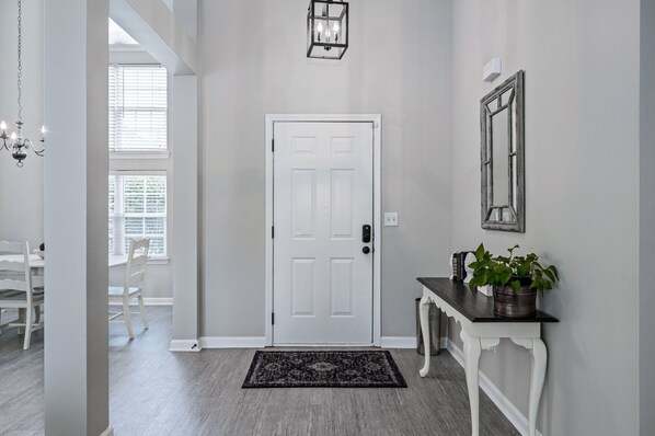 Un couloir avec une porte blanche, un tapis noir, une console blanche ornée d’une plante et un luminaire fixé au mur.