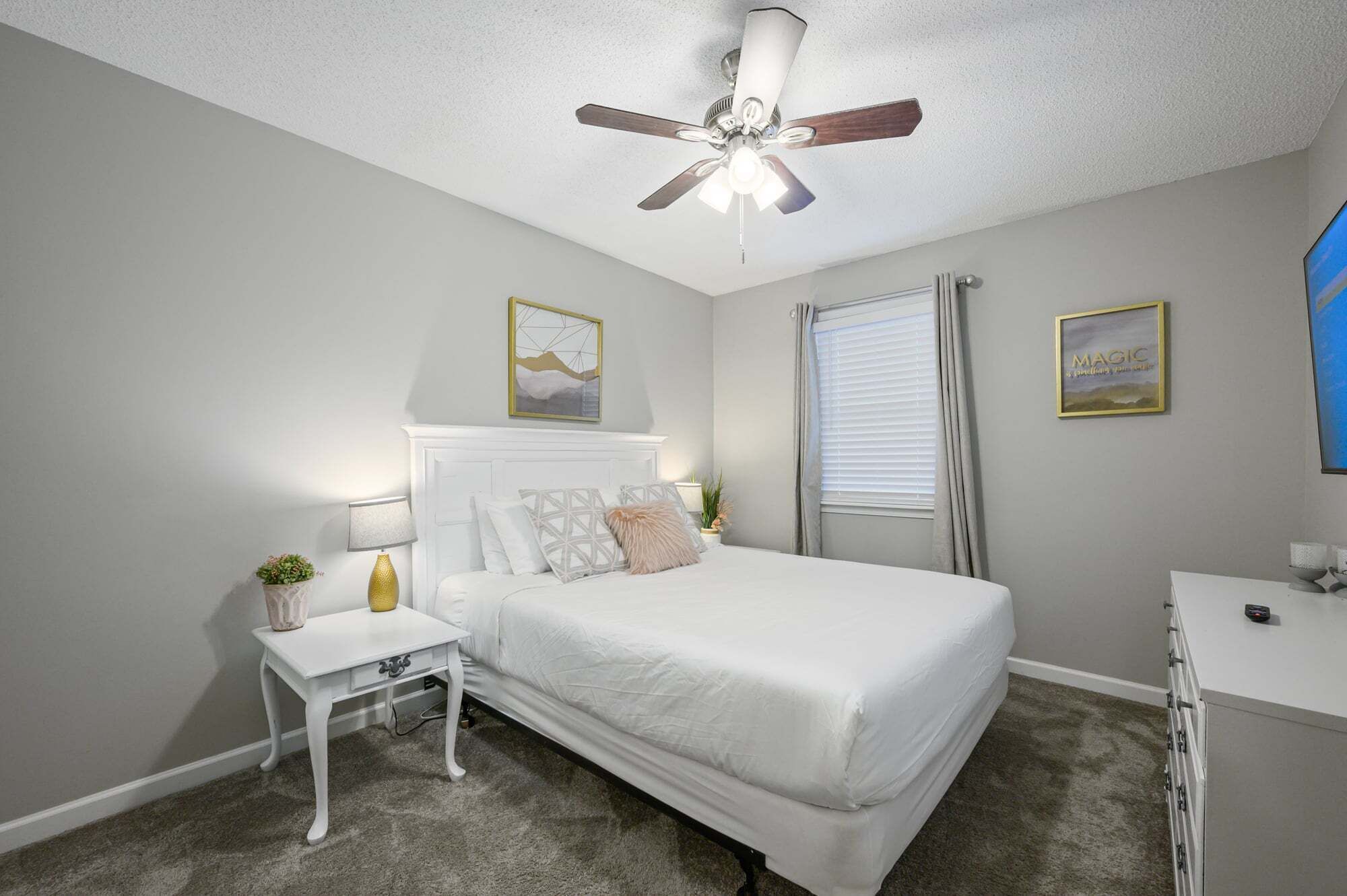 A bedroom with a bed, a nightstand, a desk, a ceiling fan, and a window with blinds.
