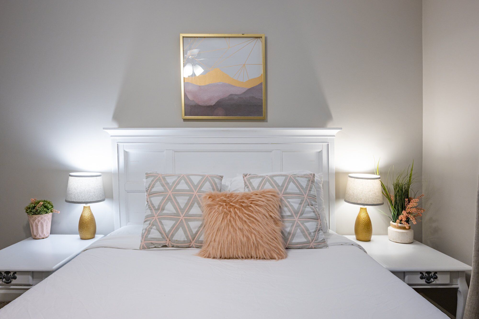A bedroom with a white bed, geometric patterned pillows, a fluffy accent pillow, bedside tables with lamps, and a framed artwork above the bed.