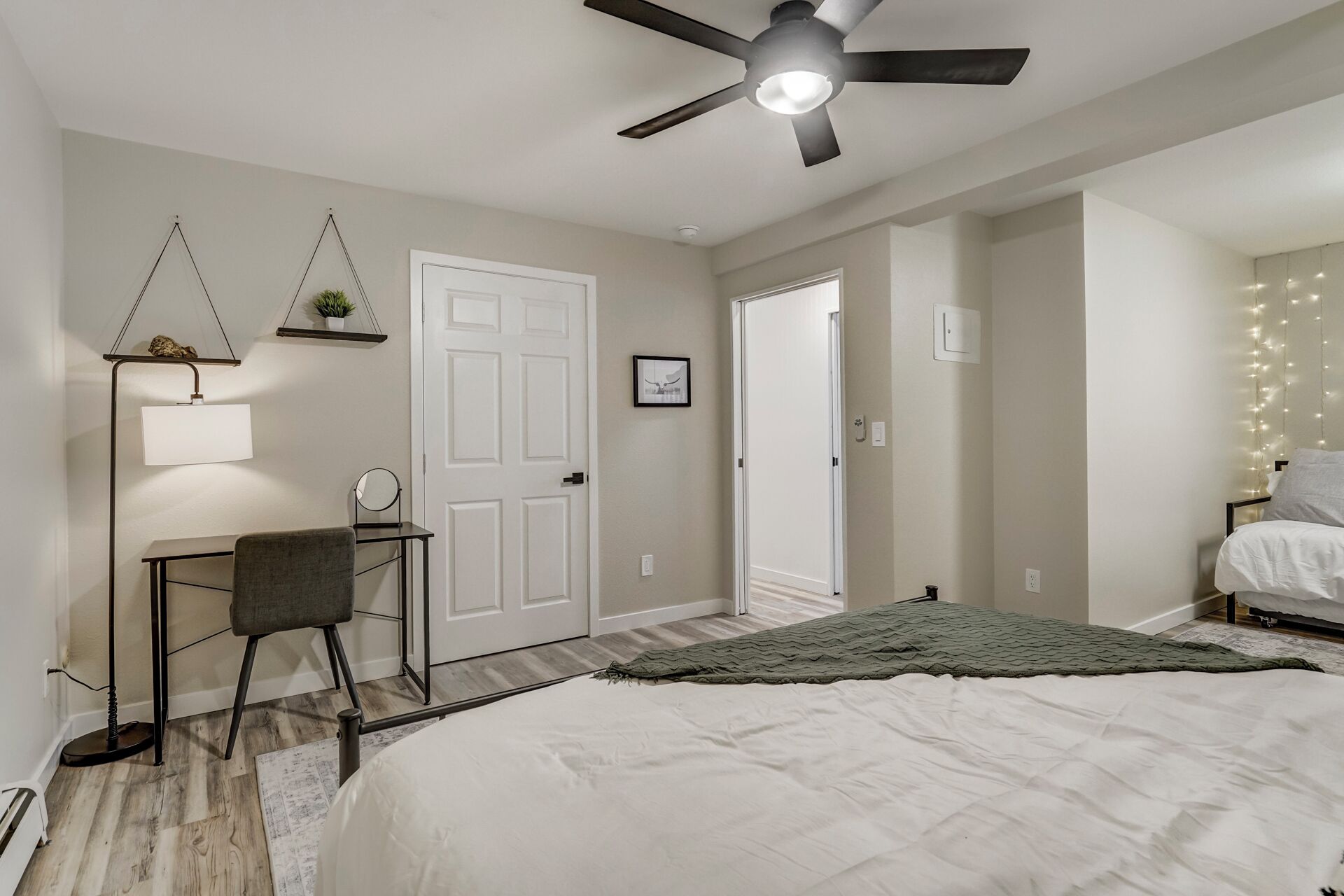 A bedroom with a bed, a desk with a chair, a ceiling fan, and a door leading to another room.