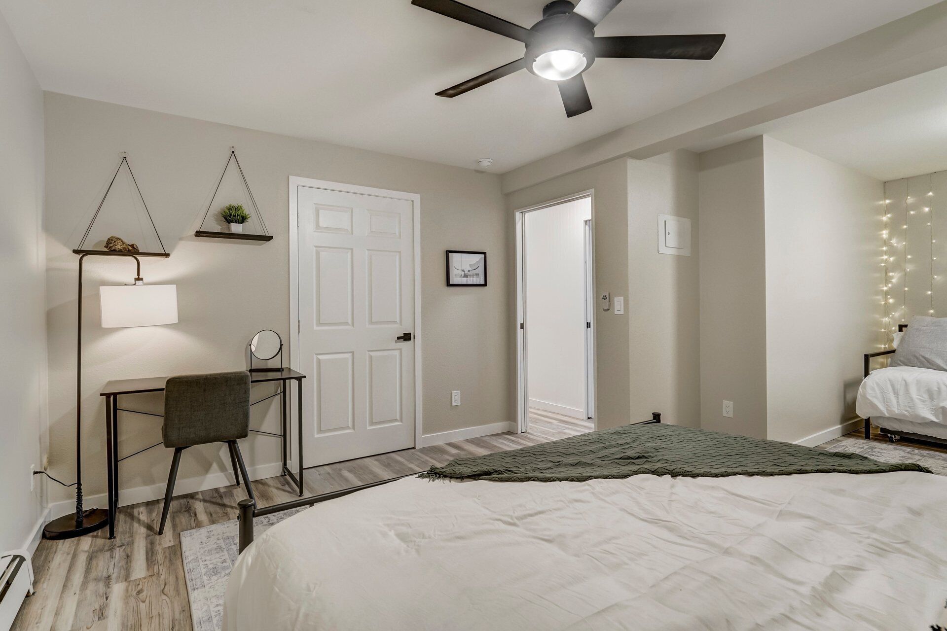 A bedroom with a bed, a desk with a chair, a ceiling fan, and a door leading to another room.