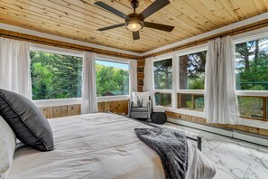 A bedroom with a large bed, a ceiling fan, and multiple windows offering a view of the forest. - 4925 Pike (Cascade)