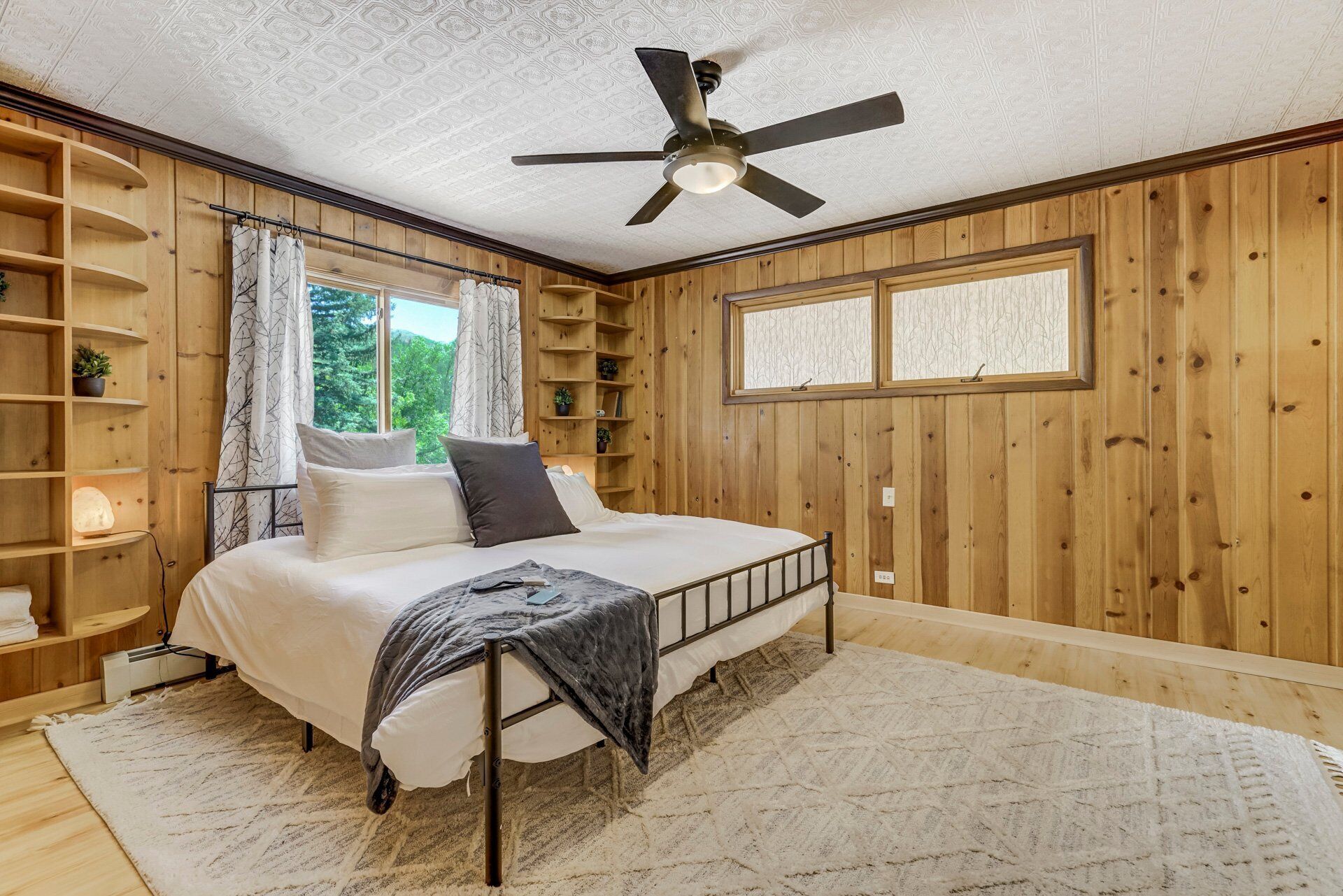 Un dormitorio con cama, ventilador de techo, paredes de madera, ventana y estanterías empotradas.