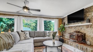 A living room with a sectional sofa, a round coffee table, a stone fireplace, and large windows with curtains. - 4925 Pike (Cascade)