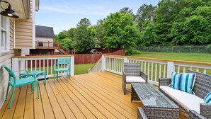 Une terrasse en bois, dotée de mobilier d’extérieur, d’une rambarde blanche et offrant une vue sur un jardin avec des arbres et une clôture.