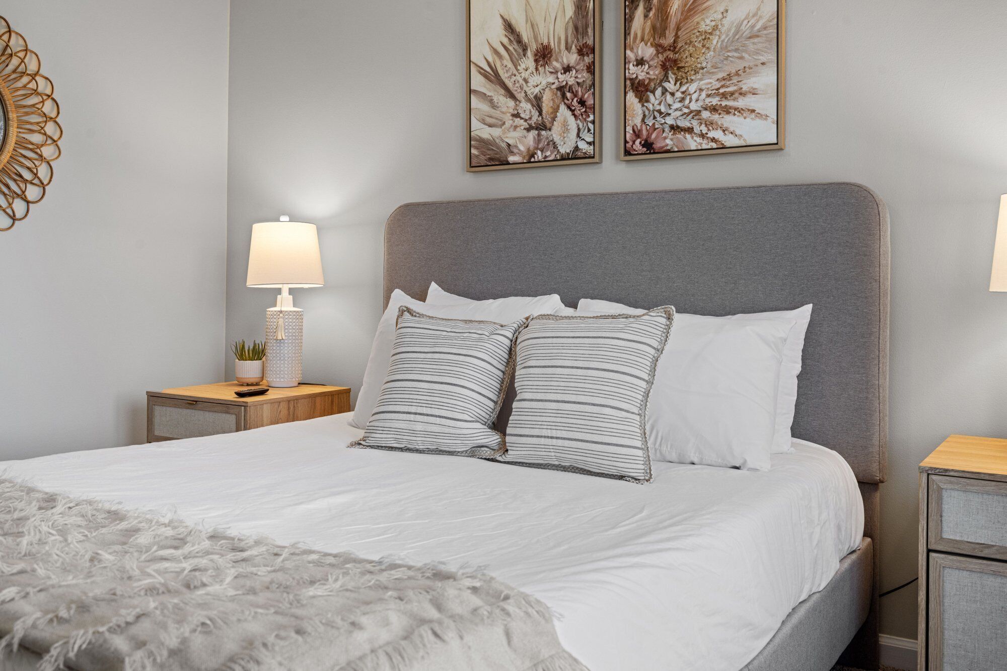 A neatly made bed with white bedding and striped pillows, a grey upholstered headboard, and two wall-mounted floral artwork pieces.