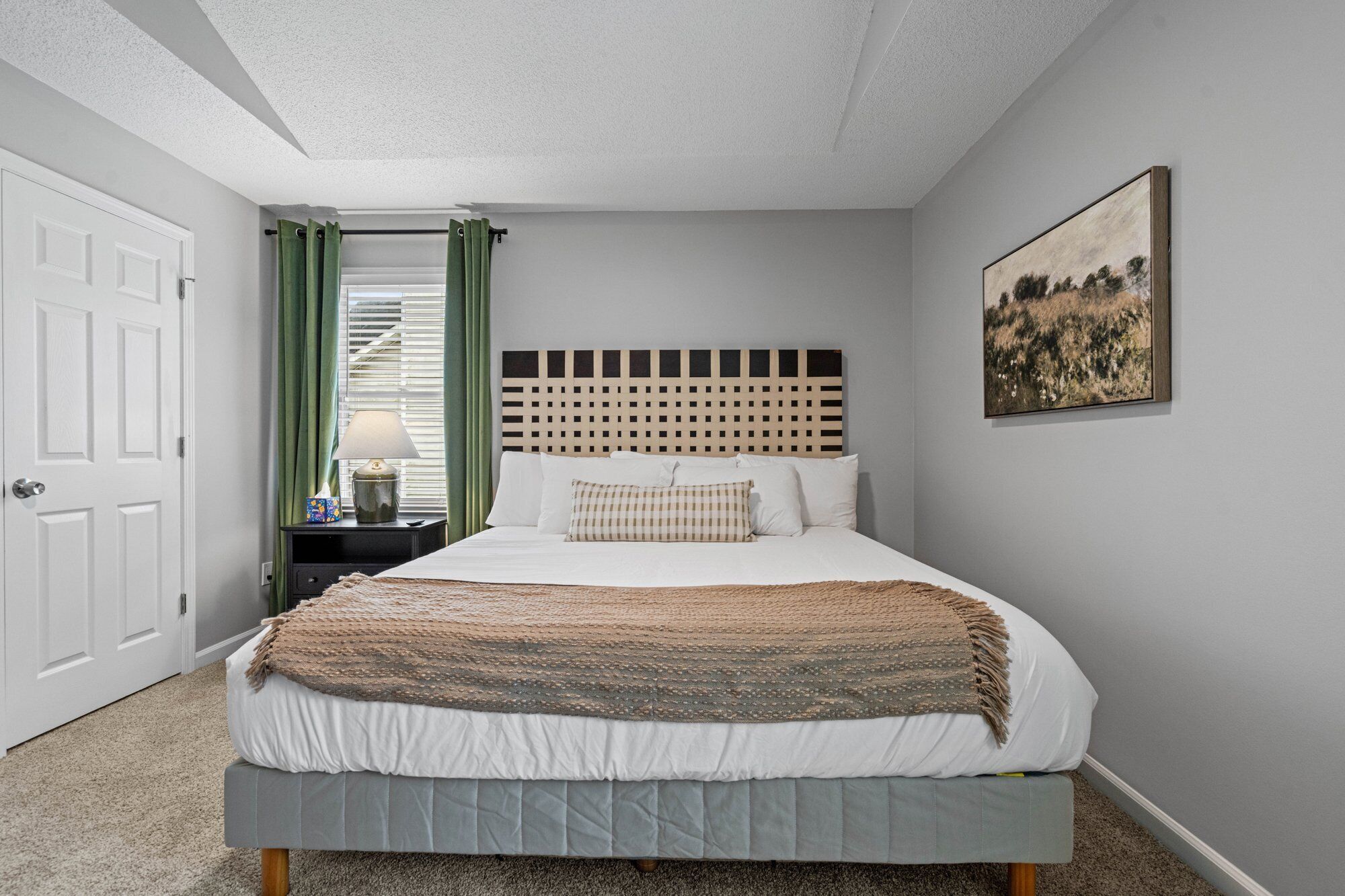 A bedroom with a bed, a headboard, a nightstand, a lamp, a door, and a painting on the wall.