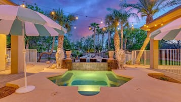 A swimming pool with a hot tub, surrounded by palm trees and outdoor umbrellas.