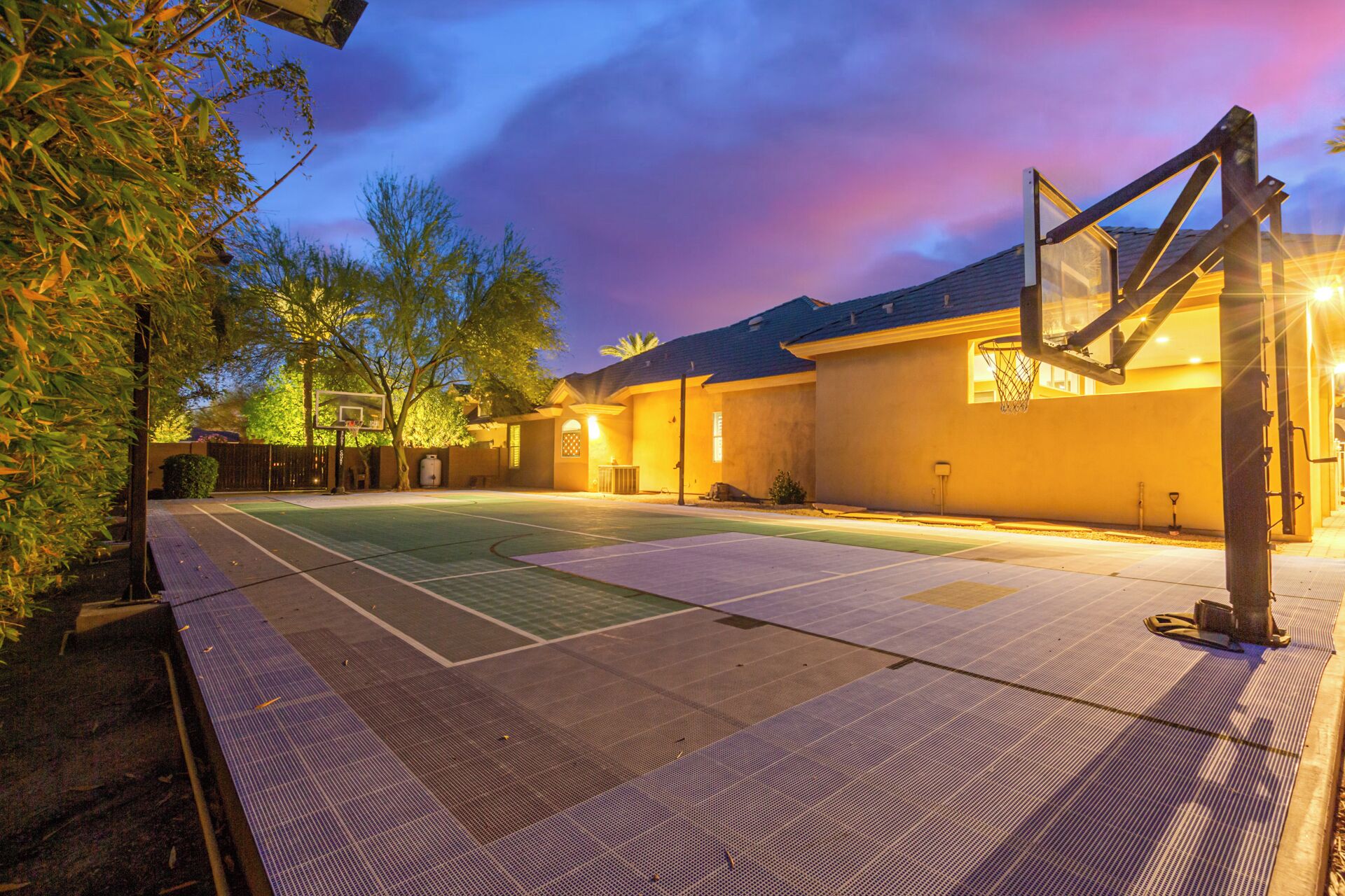 A basketball court with a hoop, surrounded by a fence and trees, under a twilight sky.