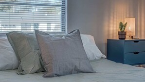 Una cama bien tendida con ropa de cama a rayas, una mesita de noche con lámpara y planta en maceta, y una ventana con persianas.