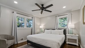 Ein Schlafzimmer mit einem Bett, zwei Nachttischen, einem Sessel, einem Deckenventilator und Fenstern mit Vorhängen.