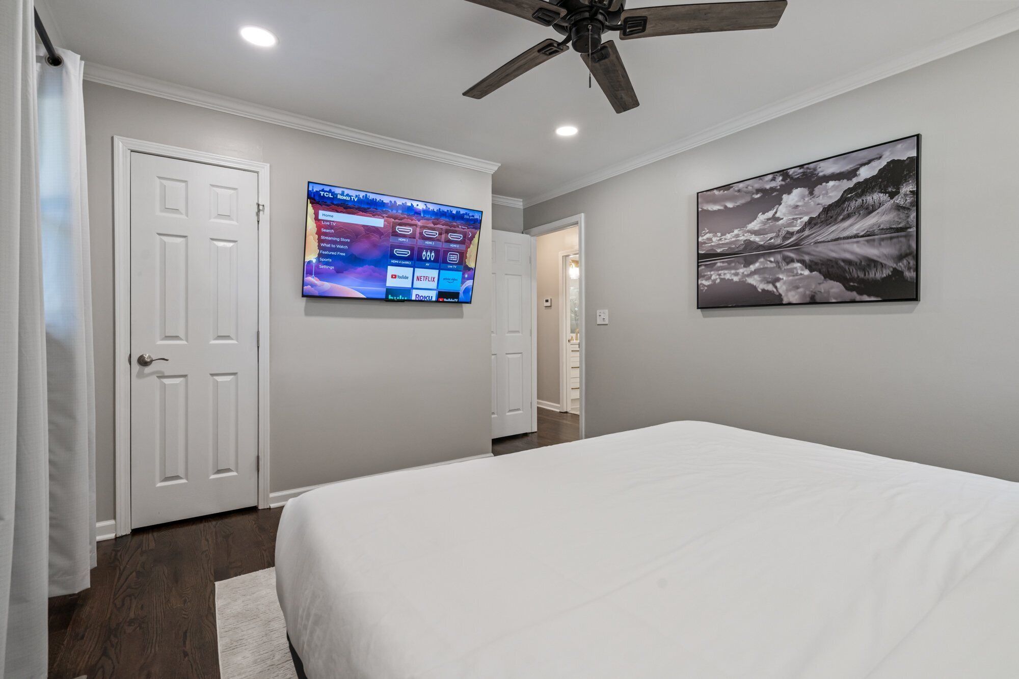 A bedroom with a bed, a ceiling fan, a mounted TV displaying a streaming service interface, and a framed picture on the wall.