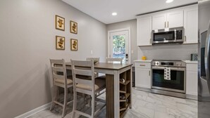 Eine kompakte Küche mit weißen Schränken, einer Mikrowelle, einem Backofen und einem Kühlschrank. Vier Stühle stehen um einen Holztisch, und eine Tür führt zu einem Balkon.