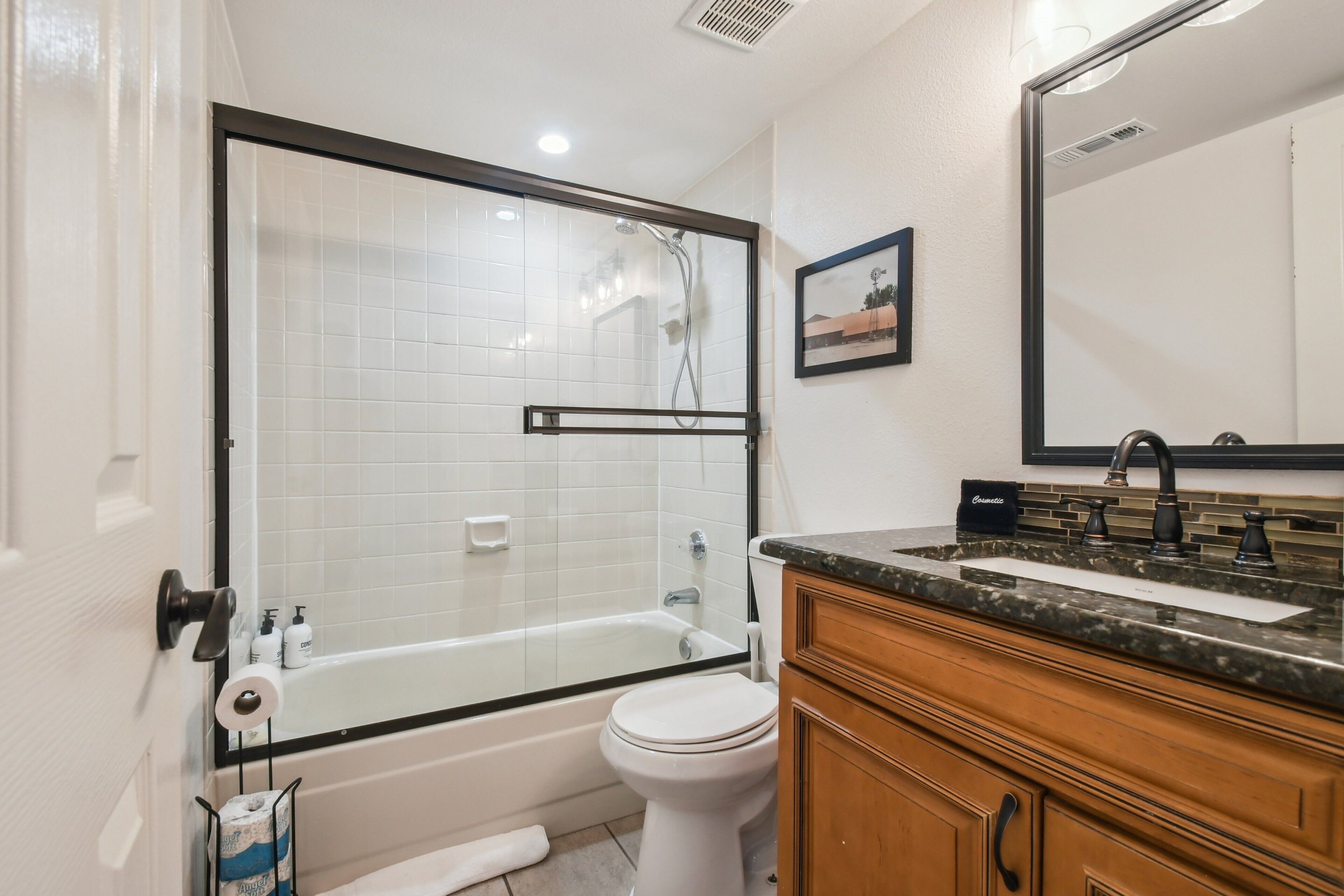 Un baño con ducha envuelta en vidrio, inodoro blanco, lavamanos con encimera de granito y muebles de madera.