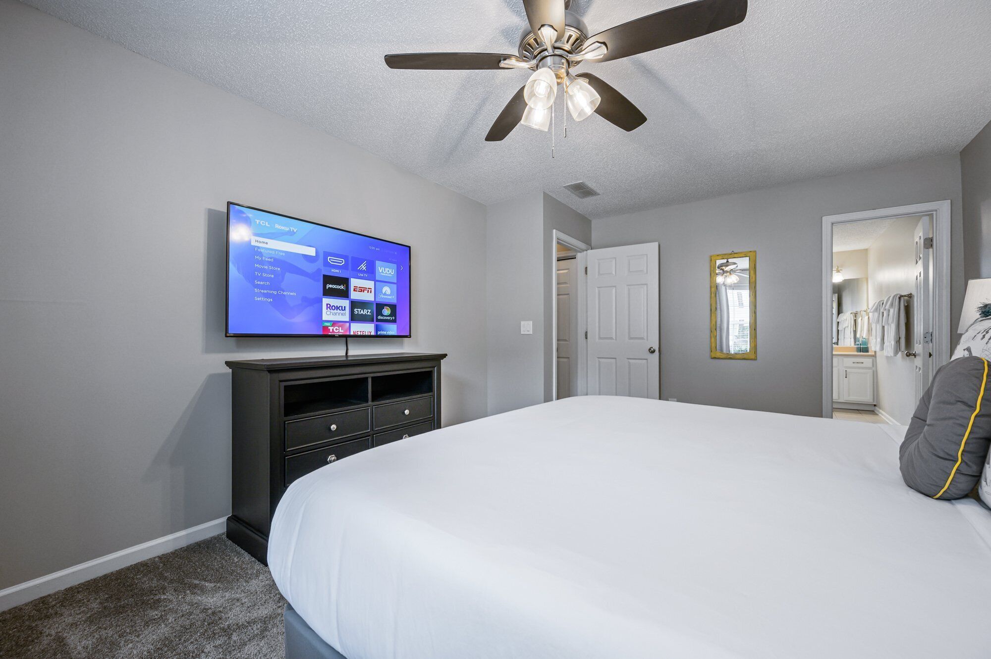 A bedroom with a bed, a TV mounted on the wall, a ceiling fan, and a door leading to a bathroom.