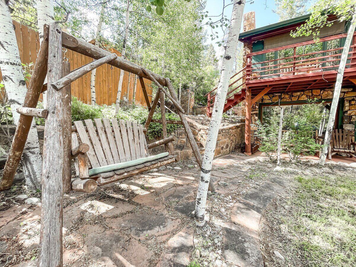 A rustic wooden swing set in a garden area with a wooden bench and a stone wall in the background.