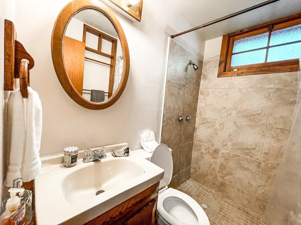 A bathroom with a wooden vanity, a round mirror, a toilet, and a shower area.