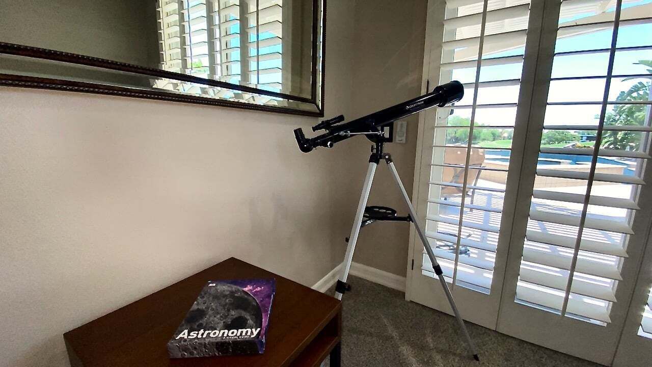 A room with a telescope on a tripod, a wooden table with a book titled 'Astronomy', and a large window with blinds.
