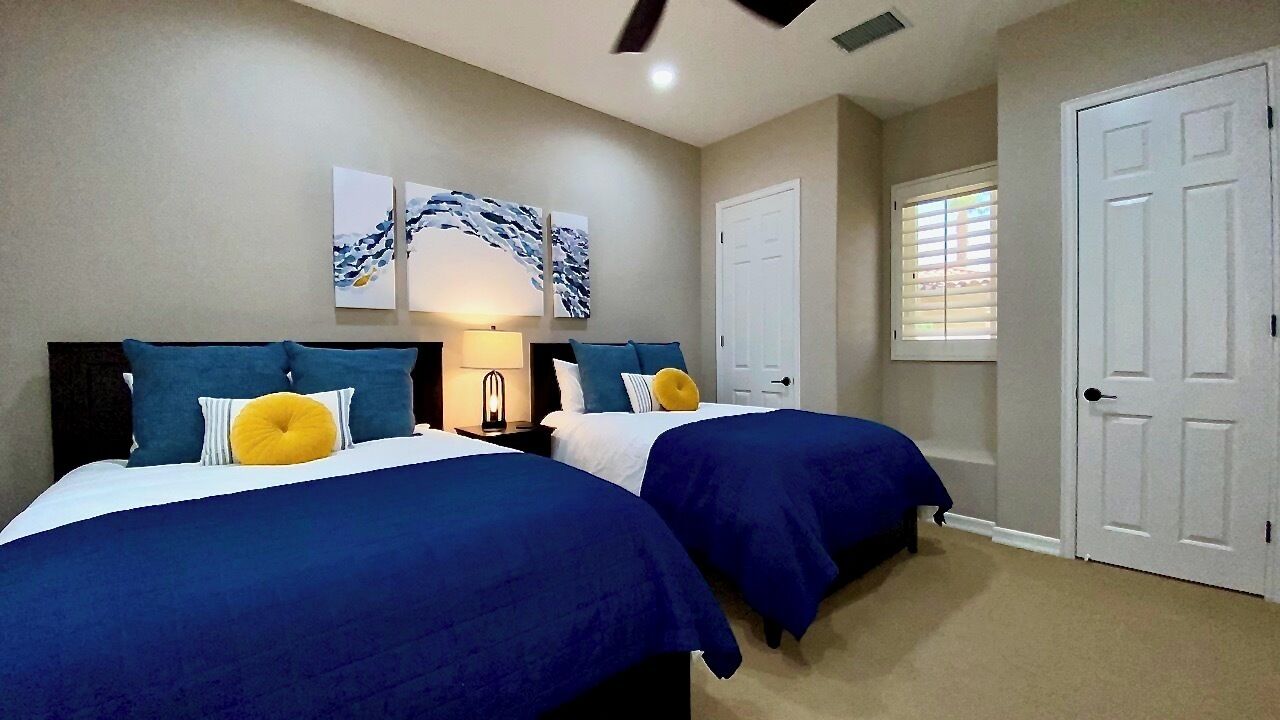 A bedroom with two beds, each with blue bedding and a yellow pillow. There are two wall-mounted lamps, a door, and a window with blinds.