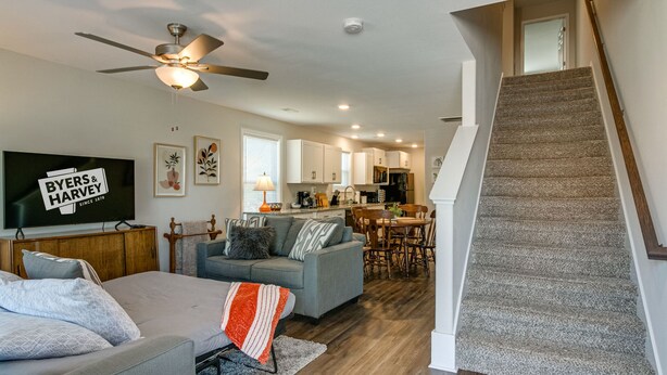 A modern living room with a grey sofa, a television, and a ceiling fan. A staircase leads to a kitchen area with white cabinets and a dining table. - Unit #A 1010 Franklin St (Clarksville)