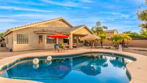 Una casa con piscina, mobiliario de patio y un paraguas de paja.