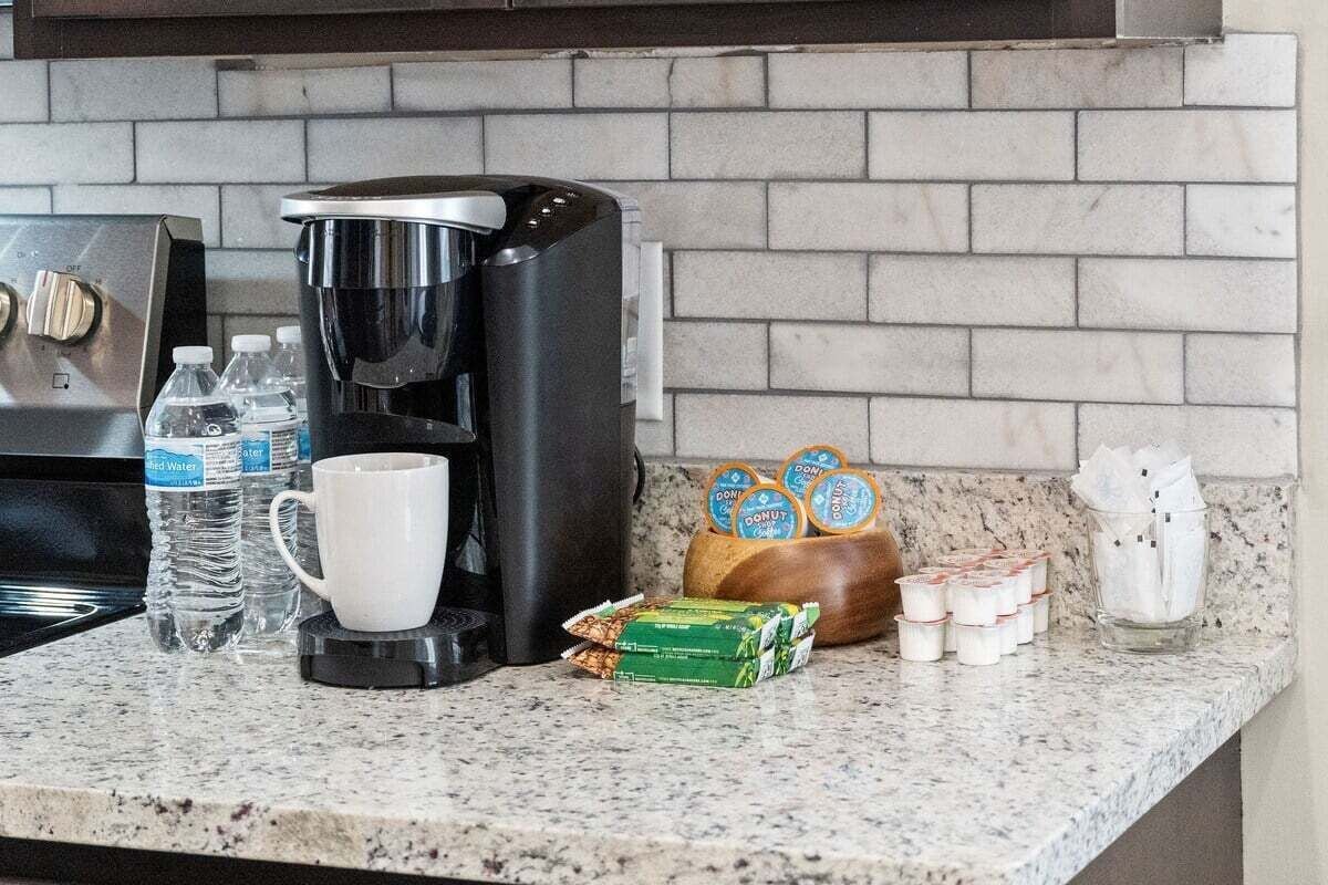 A kitchen counter with a coffee maker, water bottles, a mug, a box of donuts, and a glass container with napkins.