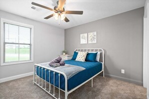 Un dormitorio con cama, ventilador de techo, ventana y dos cuadros colgados en la pared.