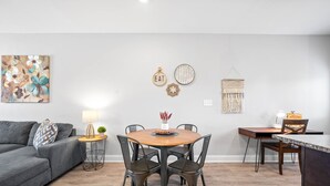 Un salon moderne avec un canapé gris, une table à manger en bois avec quatre chaises, une petite table d’appoint avec une lampe et une œuvre en macramé accrochée au mur.