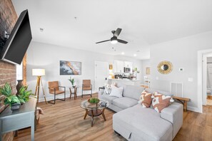 A modern living room with a grey sofa, a coffee table, and a TV on a stand. There is a kitchen in the background with white cabinets and a ceiling fan. - The Spot 281 Liberty Pkwy (Clarksville)