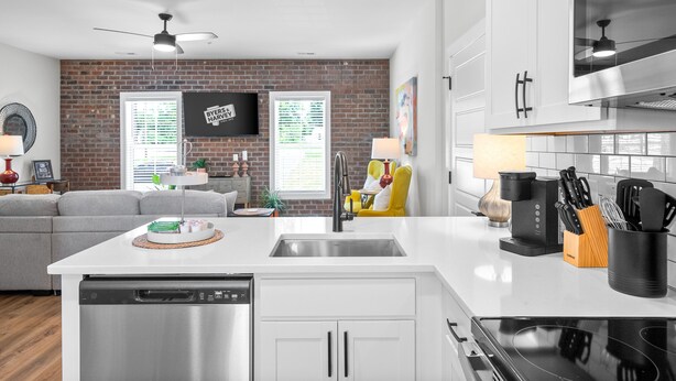 A modern kitchen with a dishwasher, sink, and stove. There is a dining area with a sofa and a table. A TV is mounted on the brick wall. - The Spot 281 Liberty Pkwy (Clarksville)