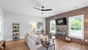 A spacious living room with a brick wall, a ceiling fan, a flat-screen TV, and a large window with blinds. - The Spot 281 Liberty Pkwy (Clarksville)