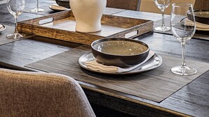 Una mesa de comedor dispuesta con platos, cuencos y copas de vino, con un florero de follaje oscuro en el centro.