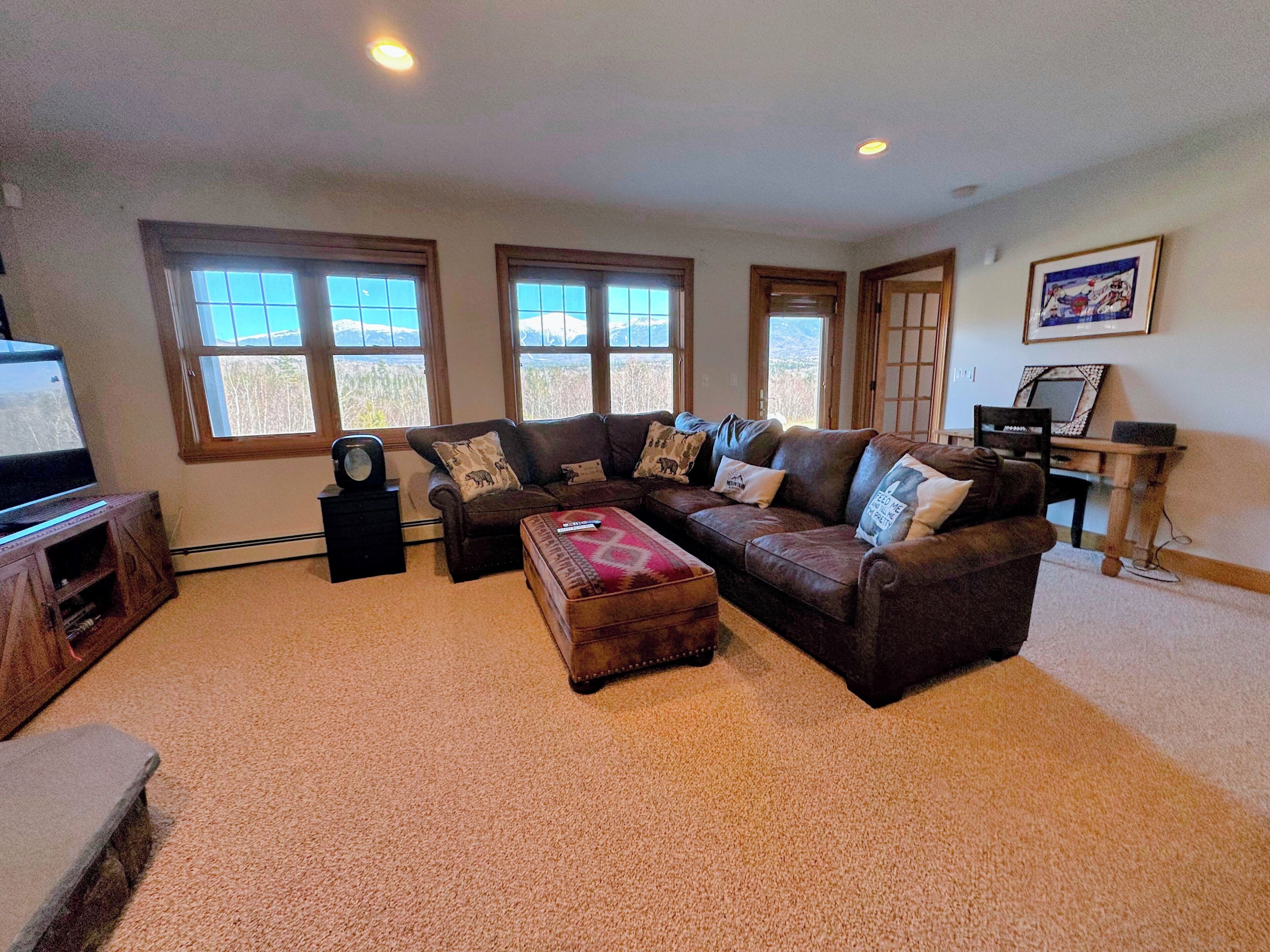 A living room with a sectional sofa, a coffee table, and a TV on a stand. There are three windows providing a view of the outdoors.