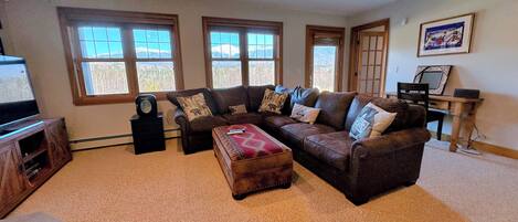 A living room with a sectional sofa, a coffee table, and a TV on a stand. There are three windows providing a view of the outdoors.
