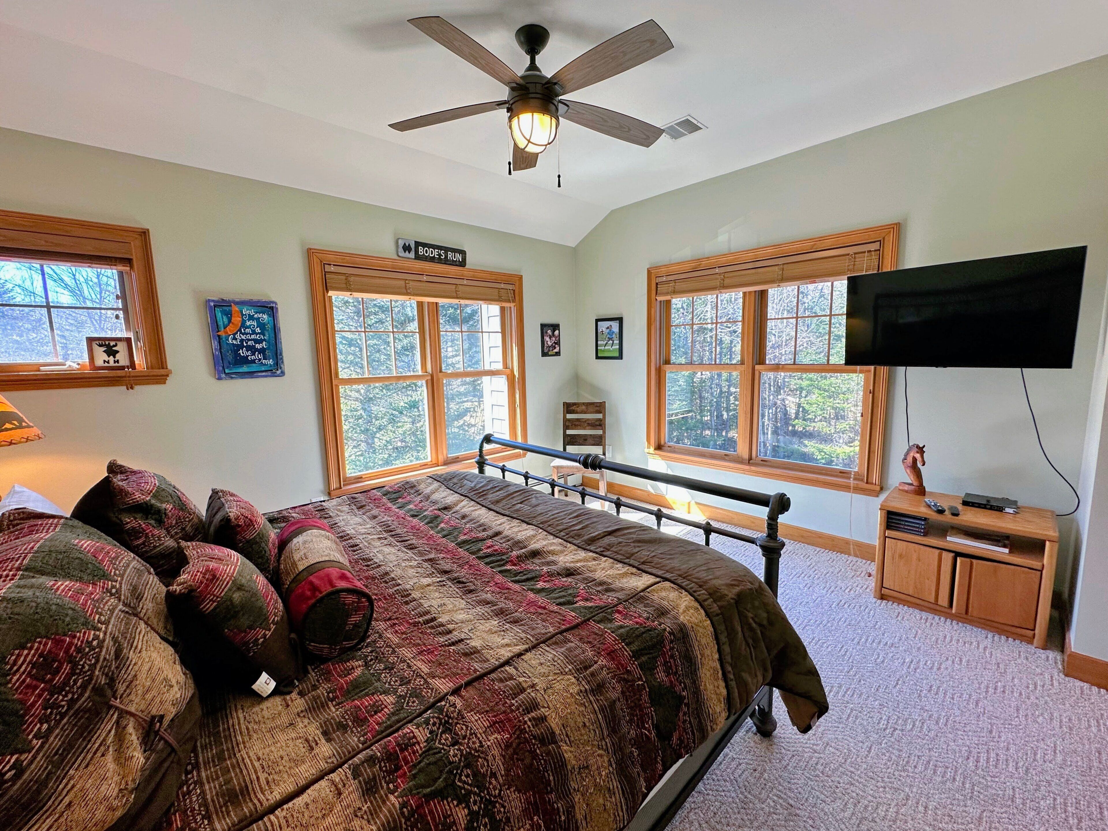 A bedroom with a bed, a ceiling fan, a TV, and large windows.